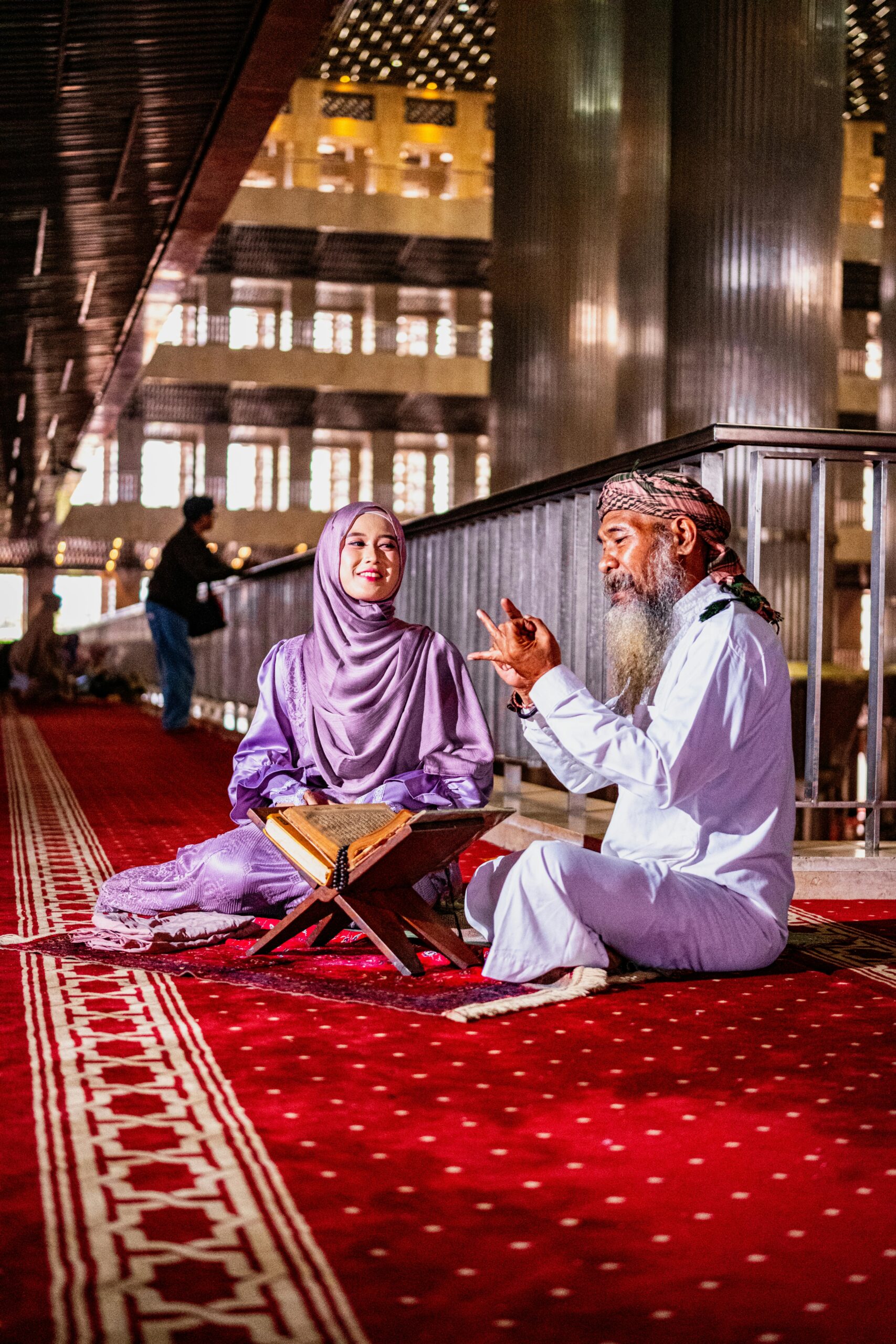 An elderly man and young woman engage in a Quran reading session in a mosque interior.
